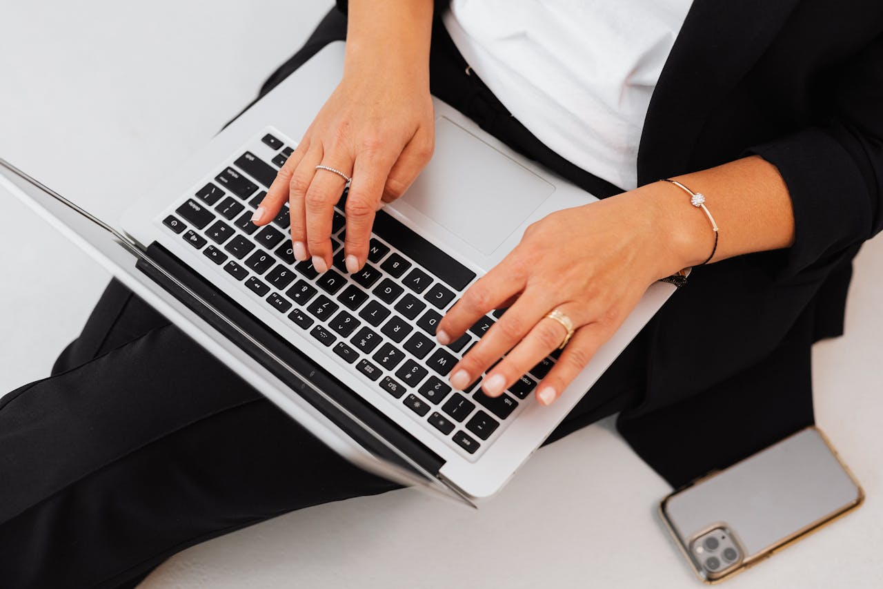 services-02 A woman in business attire types on a laptop with a smartphone beside her, emphasizing modern technology and work.